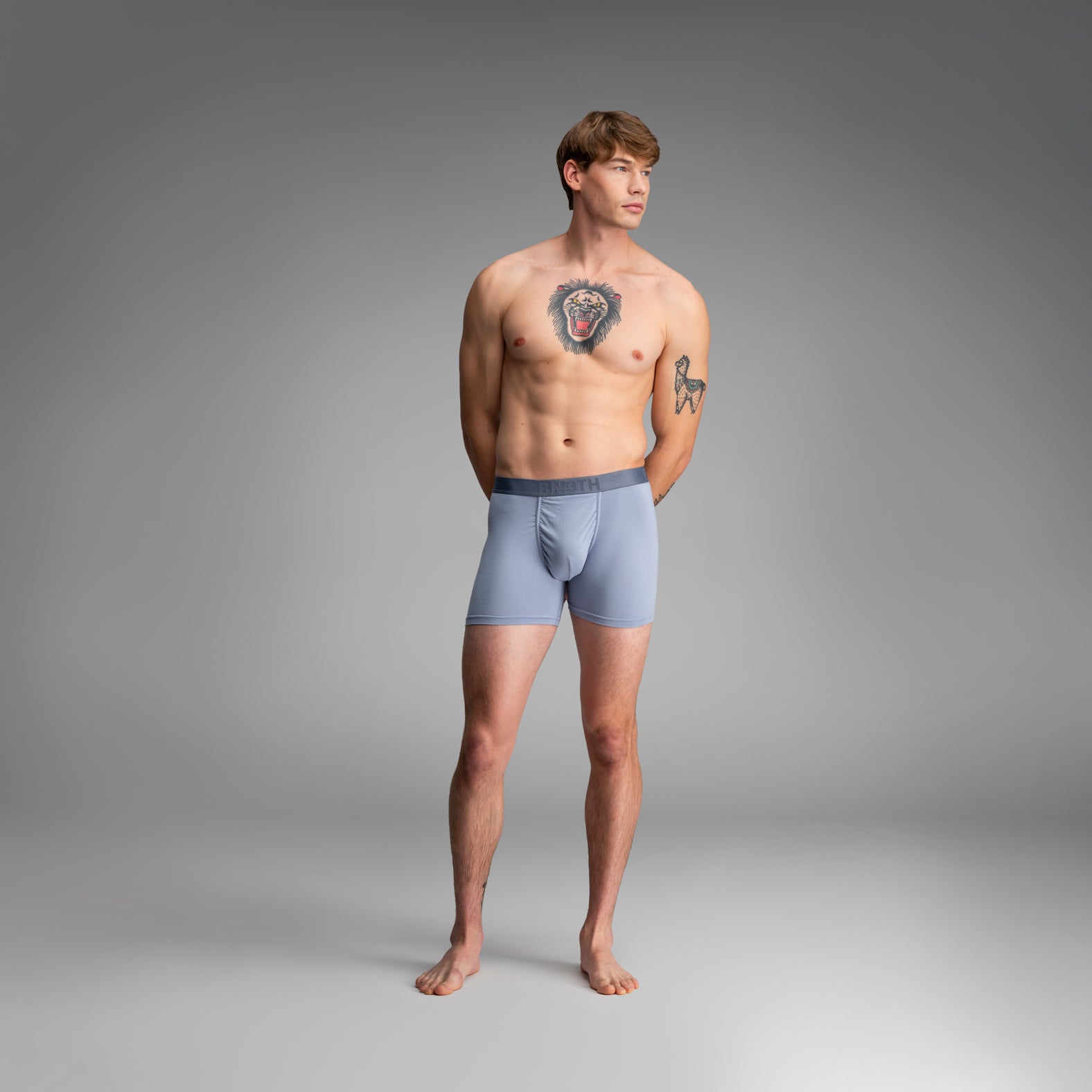 A male model stands against a smooth gray studio background wearing light blue boxer briefs with a darker gray waistband.