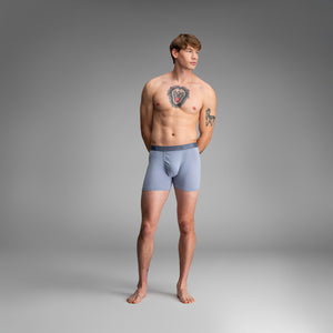 A male model stands against a smooth gray studio background wearing light blue boxer briefs with a darker gray waistband.