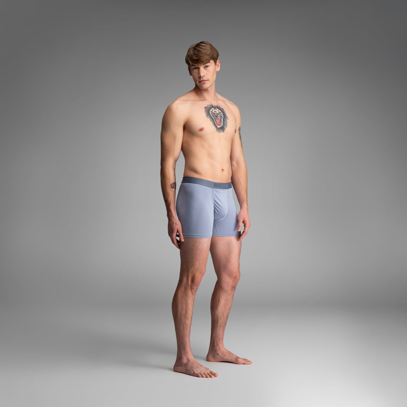 A male model stands barefoot against a smooth gray studio background, wearing light blue boxer briefs with a darker gray waistband. 
Essentials Boxer Brief: Dusty Blue
