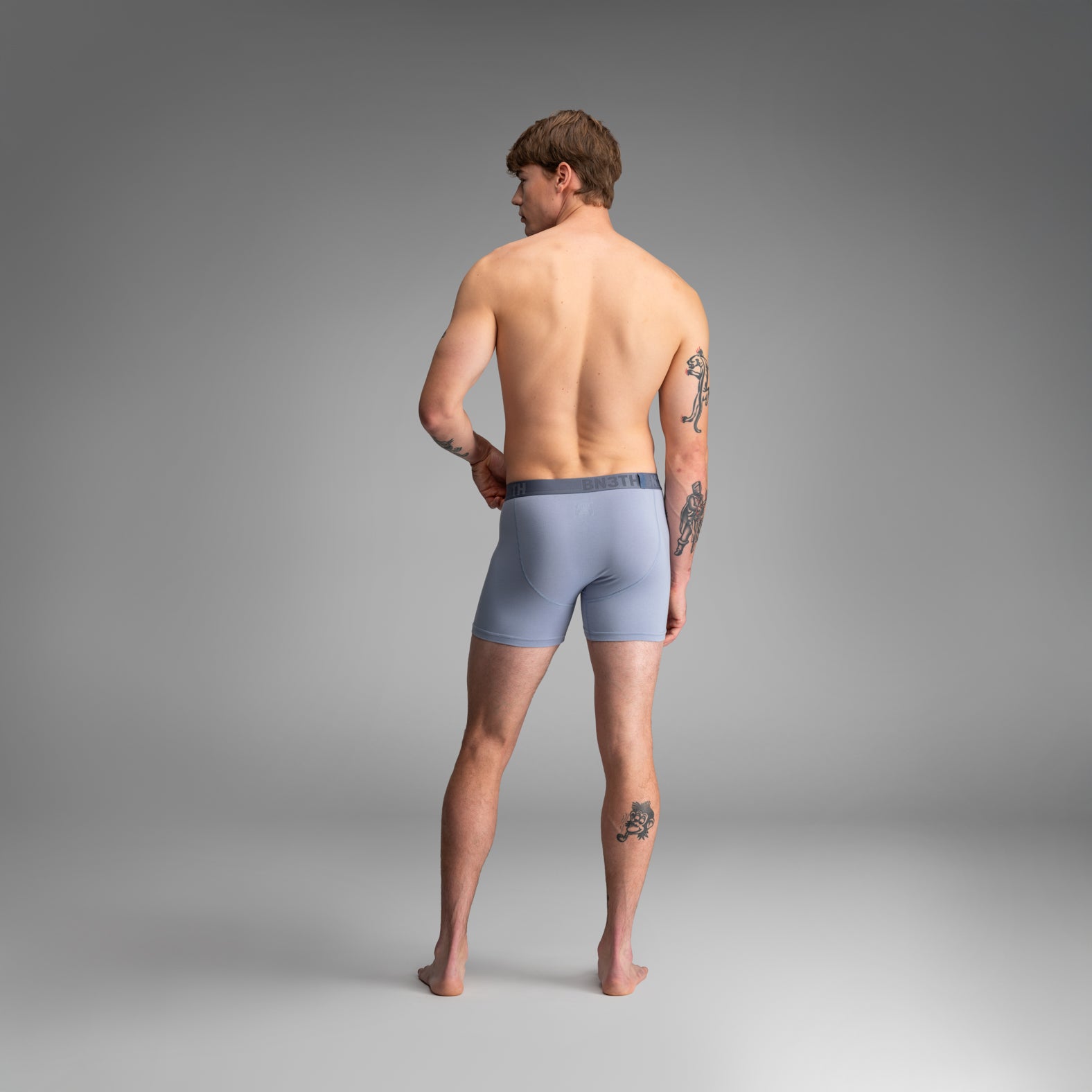 A man stands with his back to the camera wearing light blue boxer briefs against a smooth gray studio background. 
Essentials Boxer Brief: Dusty Blue
