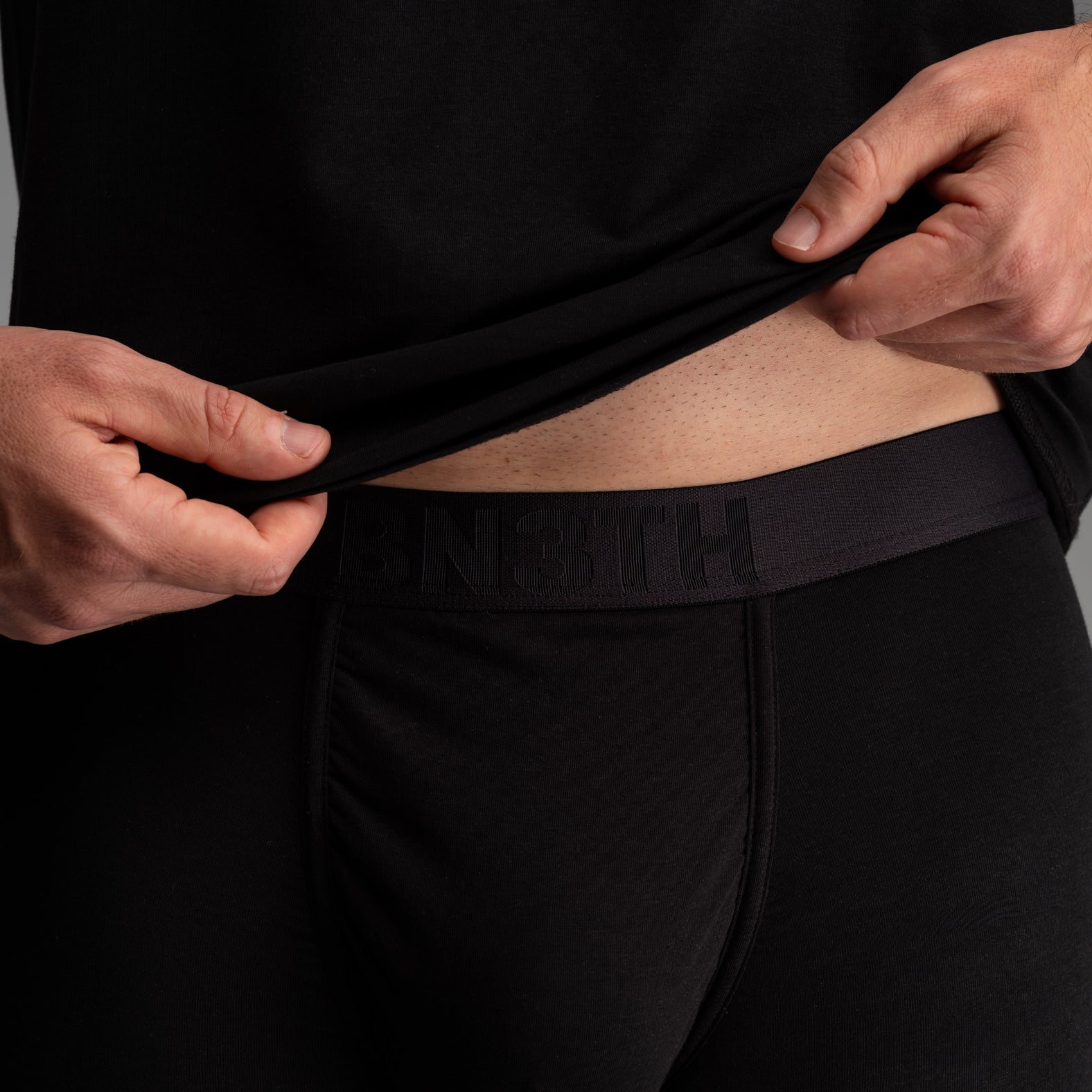 Close-up of a person wearing black underwear and lifting the hem of a black shirt to show the black branded waistband with the word 'BN3TH' embossed. Essentials Boxer Brief: Black