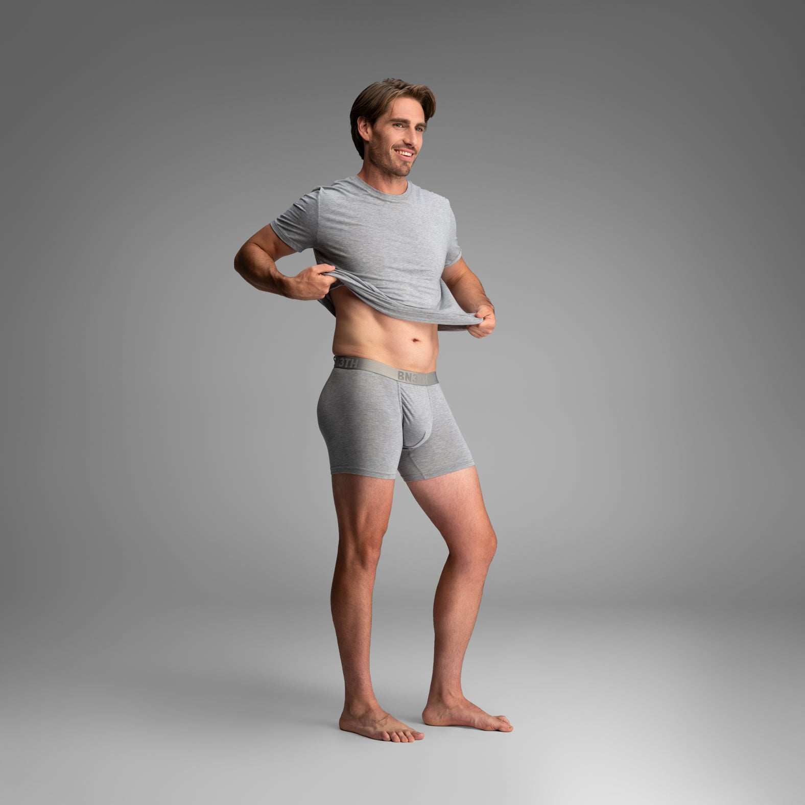 A man modeling a light gray crewneck t-shirt and matching gray boxer briefs, lifting the shirt to show fit against a neutral gray studio background .Essentials Boxer Brief: Heather Gray